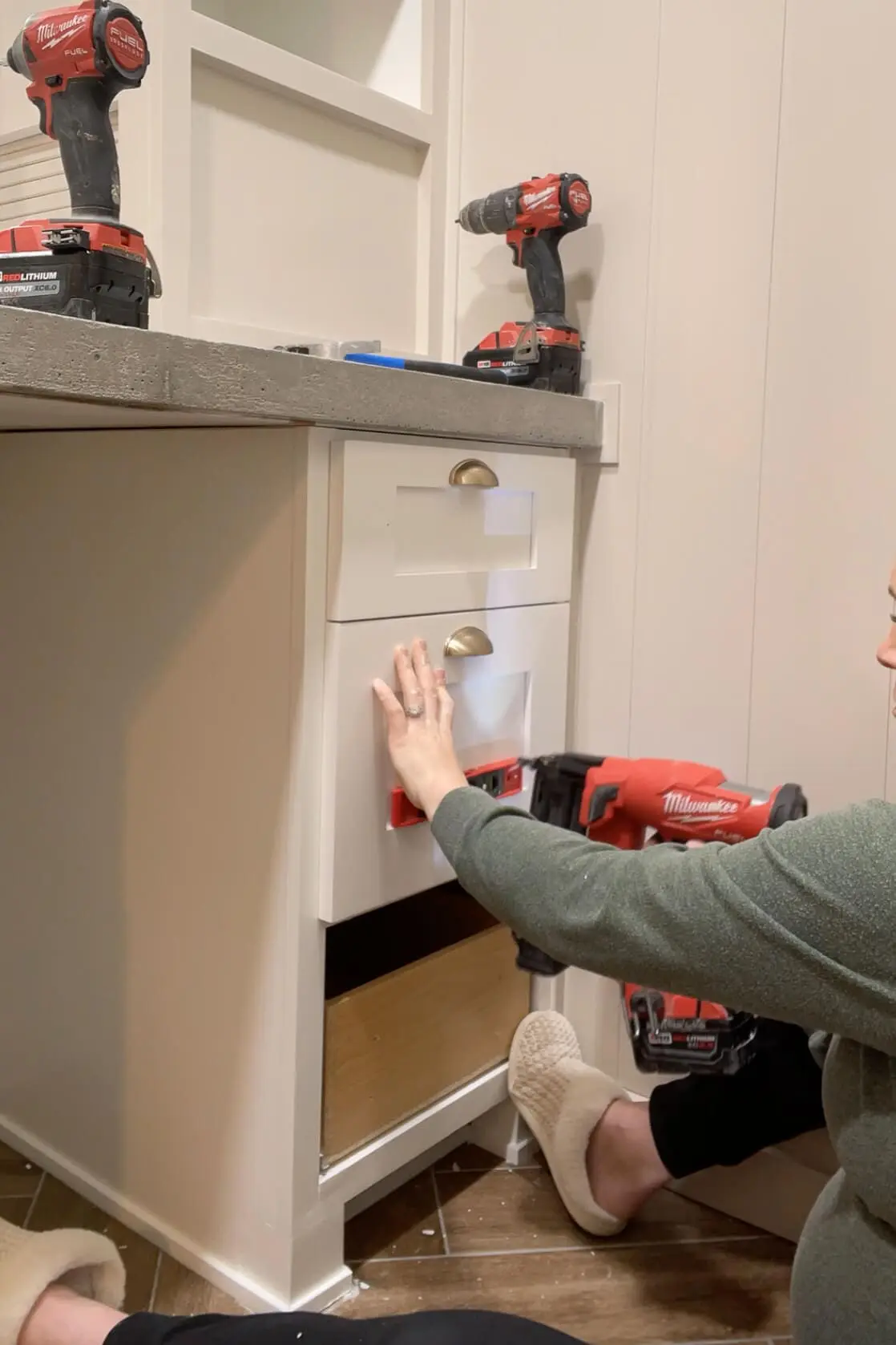 Person sitting on the floor, using a red nail gun to install and align a white drawer front on a bathroom cabinet with a concrete countertop. Power tools and a built-in shelving unit are visible in the background.